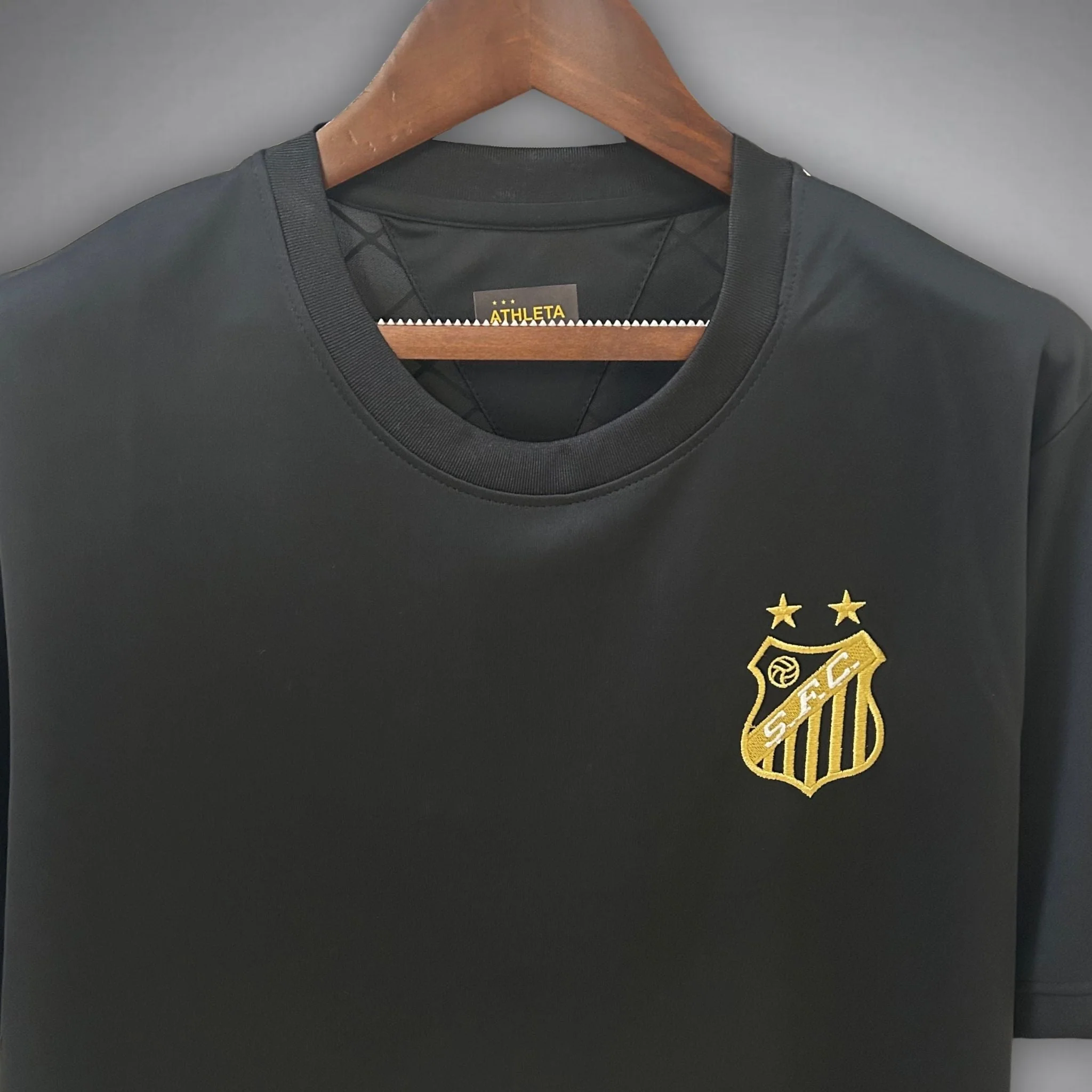Upper-front detail of the Santos FC Perola Negra black shirt, with a ribbed round neckline, Athleta neck label and gold SFC crest topped by two stars