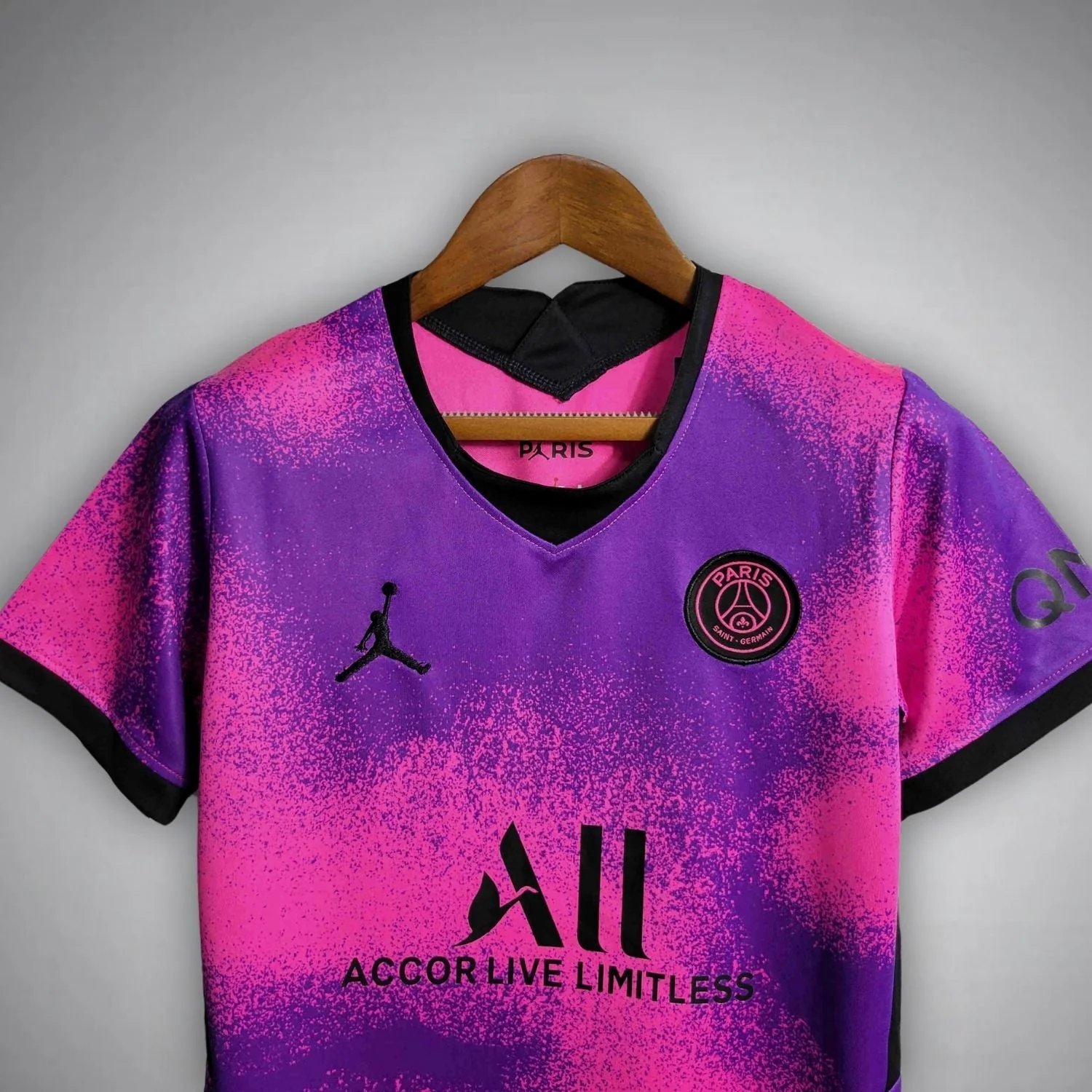 Close-up of the black neckline, PSG crest and ALL sponsor lettering on a pink-purple kids football shirt.
