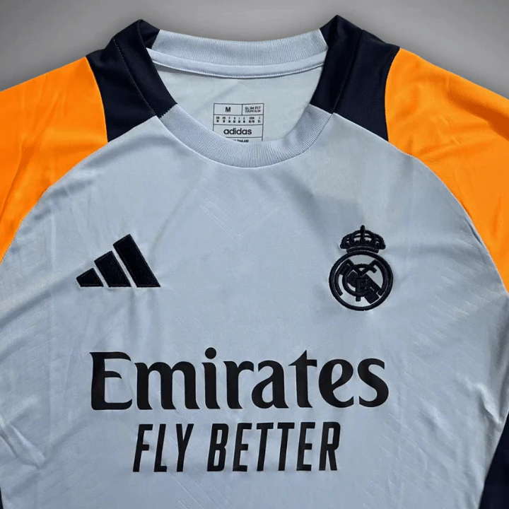 Chest and collar close-up of the Real Madrid 2024/25 pre-match shirt, with a pale grey round neckline, black crest and adidas mark, black sponsor lettering and orange shoulders
