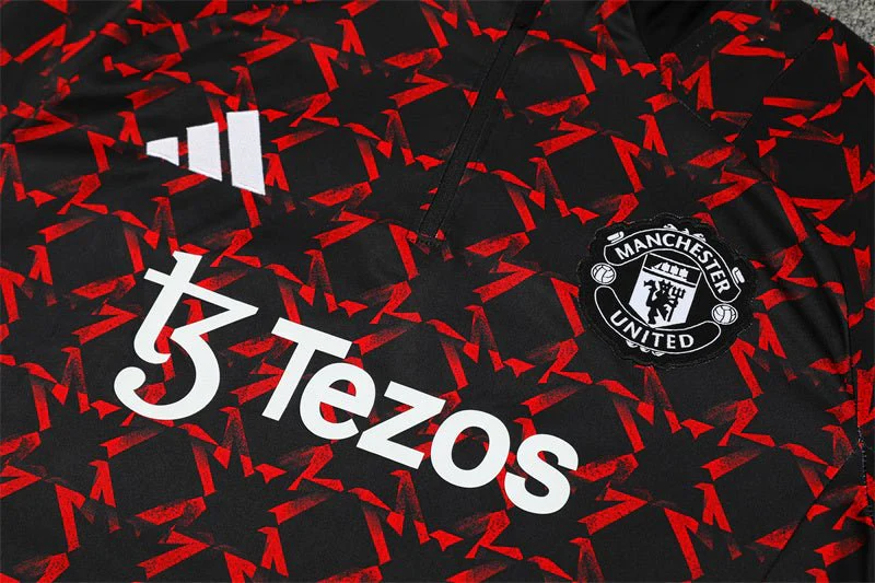 Close-up of white Tezos lettering, adidas logo and a monochrome Manchester United crest on the black tracksuit top with red angular graphics