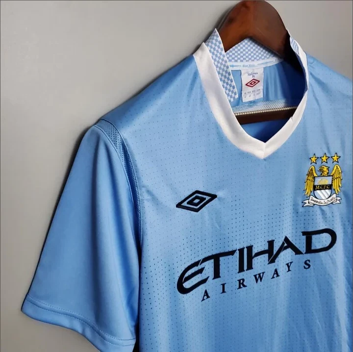 Angled chest close-up of a sky blue Manchester City retro shirt showing its white V-neck, Umbro diamond, eagle crest and ETIHAD AIRWAYS lettering.