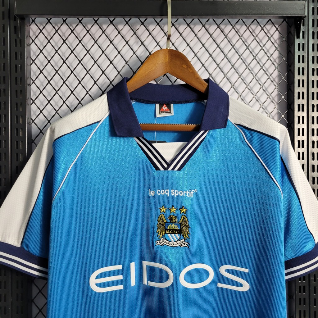 Chest detail of a sky blue Manchester City retro shirt showing a navy collar, striped V-neck, central eagle crest and white EIDOS lettering.