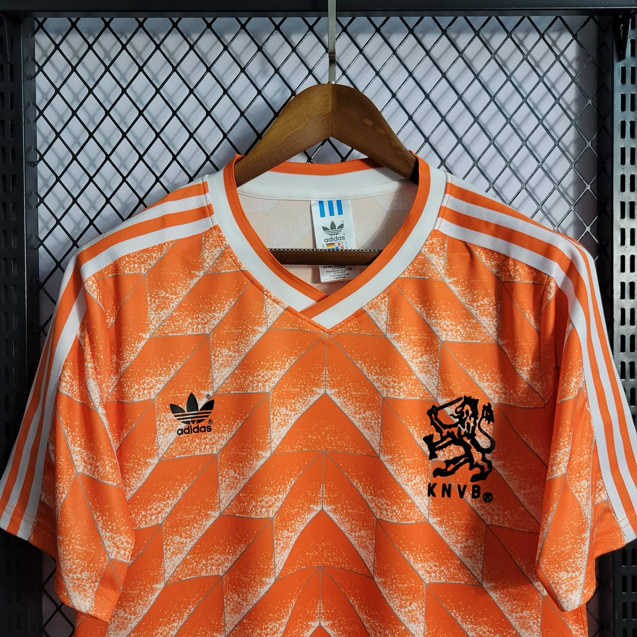 Upper-front view of the orange Netherlands shirt, showing white geometric chevrons, white V-neck and three shoulder stripes, black adidas trefoil and KNVB lion emblem